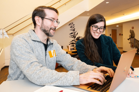 A man and woman seating at a table, the man typing on a laptop.